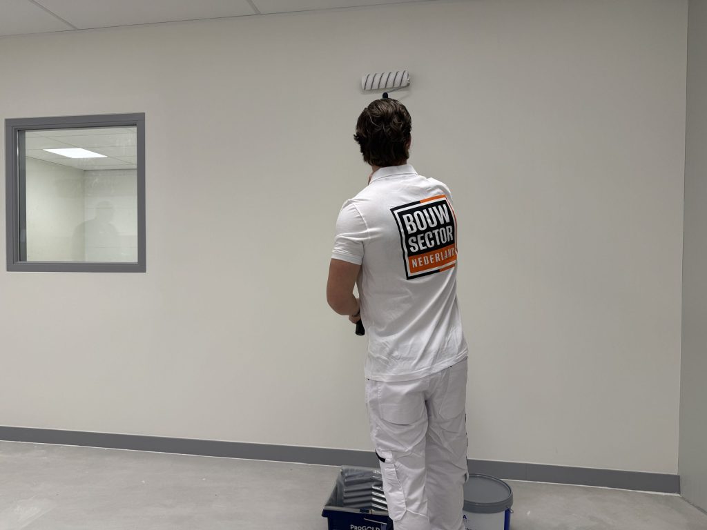 Schilder van Bouwsector Nederland staat bij zijn verfbak en werkt een witte muur af met de roller