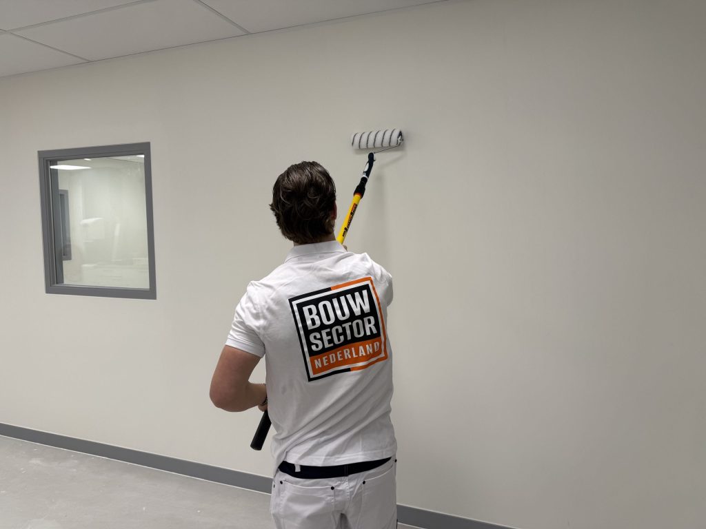 Schilder rolt een lichte wand in een gang met een grijs kozijn rond het binnenraam