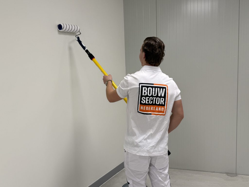 Schilder rolt een lichte wand met een roller aan een gele steel, naast een wand van metalen panelen